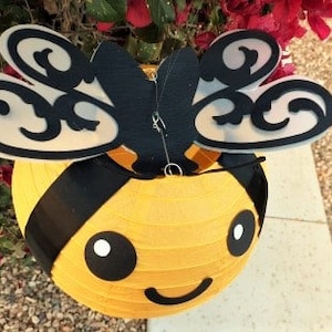 May include: A yellow and black paper lantern shaped like a bee with a smiling face. The bee has black and white eyes and black wings with a decorative pattern.