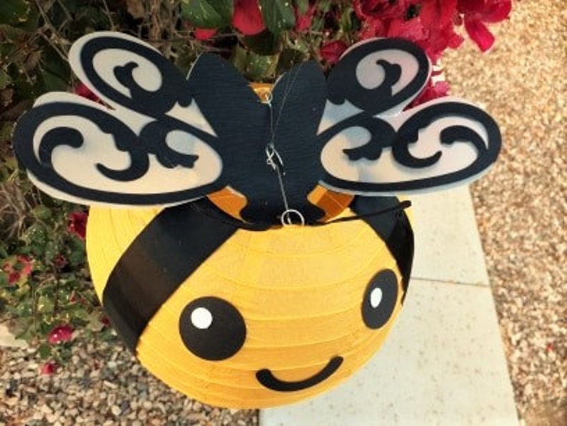May include: A yellow and black paper lantern shaped like a bee with a smiling face. The bee has black and white eyes and black wings with a decorative pattern.