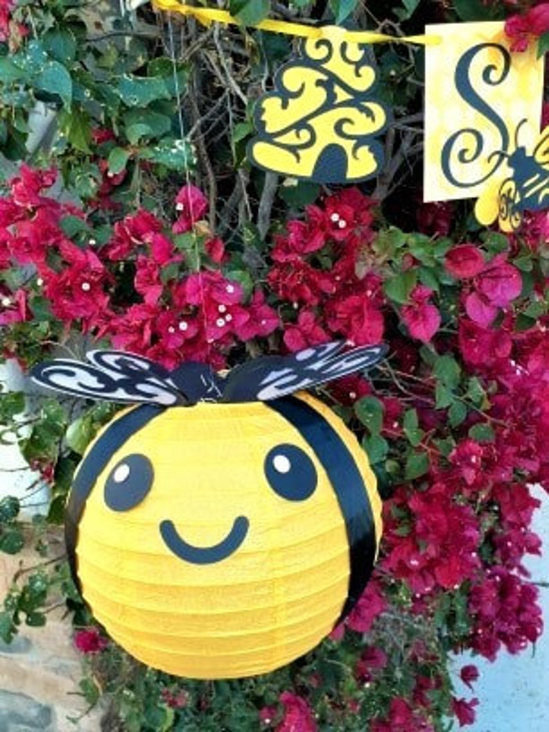 May include: A yellow paper lantern shaped like a bee with black stripes and a smiling face. The bee lantern is hanging from a branch of a bush with pink flowers.