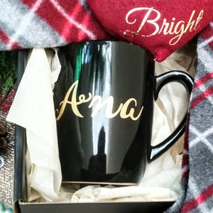 May include: A black mug with a gold inscription that says "Anna" is nestled in a white tissue paper-lined box. A red heart-shaped ornament with the text "Merry & Bright" is visible in the background.