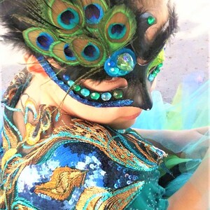 May include: A young person wearing a teal sequined costume with a peacock feather eye mask. The mask has blue and green iridescent feathers and rhinestones.