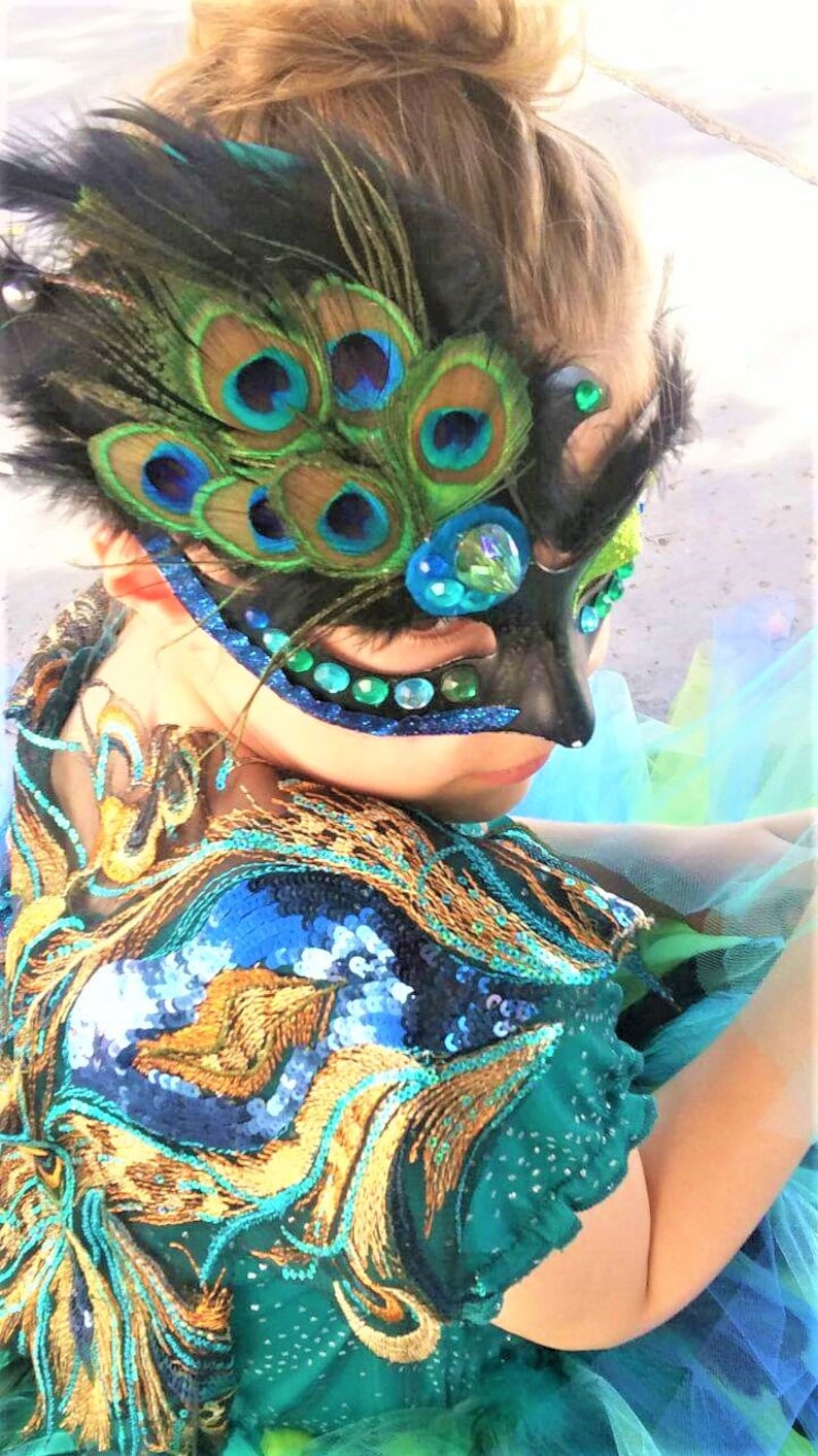 May include: A young person wearing a teal sequined costume with a peacock feather eye mask. The mask has blue and green iridescent feathers and rhinestones.