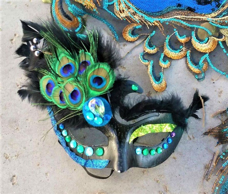 May include: A black masquerade mask with peacock feathers, green and blue iridescent sequins, and a crystal embellishment. The mask has a long, thin nose and a wide, open mouth.