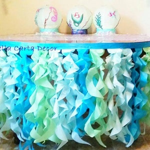 Puede incluir: Una mesa cubierta con un mantel de tela azul y verde con volantes. La mesa está decorada con tres globos de vidrio con diseños de caballitos de mar, sirenas y flores. El texto "Della Carta Decor" es visible en el mantel.