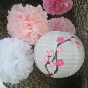 May include: White and pink paper pom-poms and a white paper lantern with pink cherry blossom decorations hanging from a tree branch.