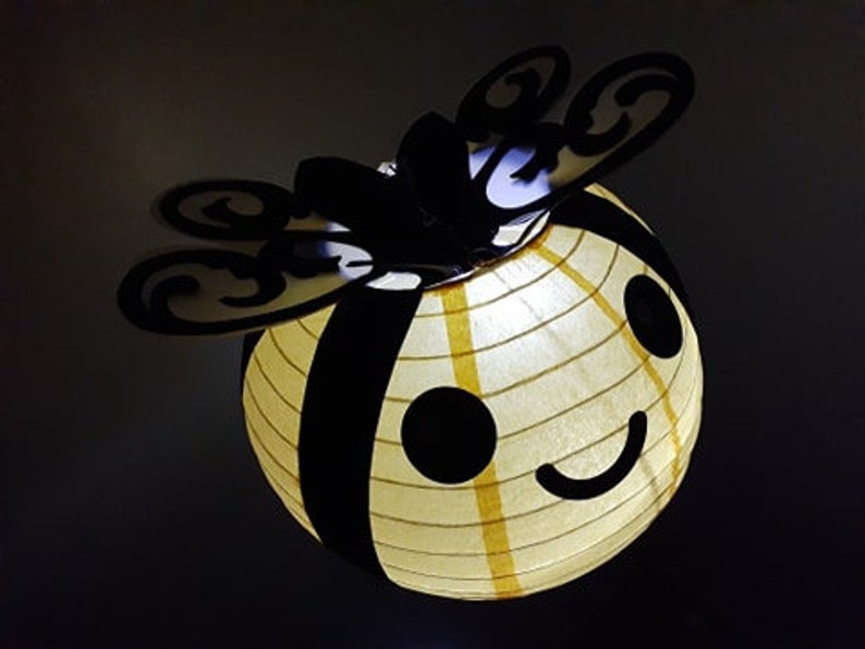 May include: A yellow and black bee-shaped paper lantern with a smiling face and black eyes. The lantern has a white paper lining and is lit from within.