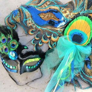 May include: A black masquerade mask with green and blue accents and peacock feathers. A peacock feather wand with a blue tulle bow.