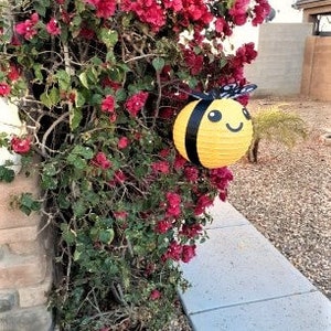 May include: A yellow and black bee-shaped paper lantern hanging from a bush of pink flowers. The lantern has a smiling face and black wings.