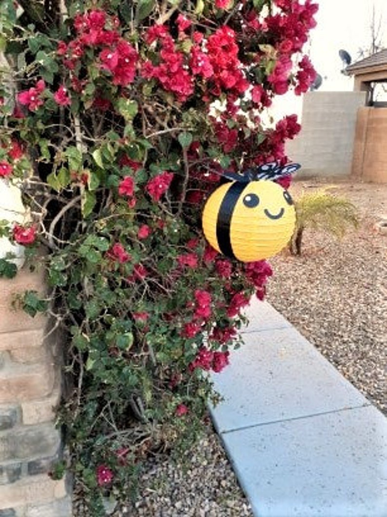 May include: A yellow and black bee-shaped paper lantern hanging from a bush of pink flowers. The lantern has a smiling face and black wings.