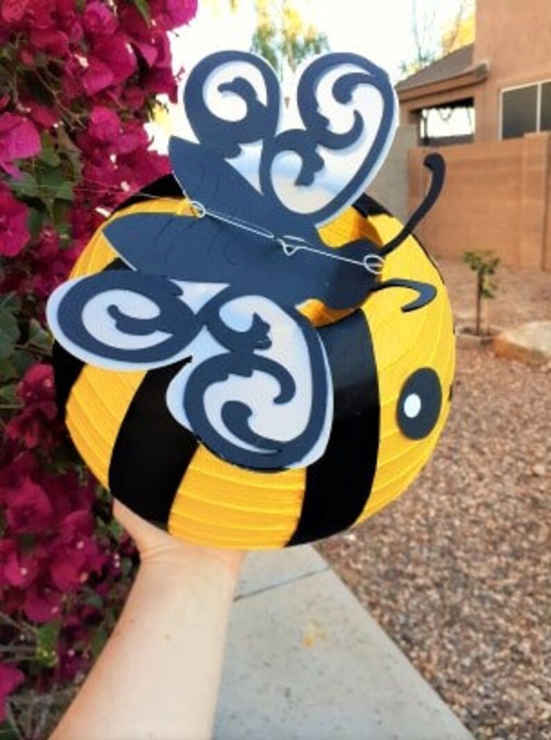 May include: A yellow and black bee-shaped paper lantern with a black butterfly cutout design. The lantern is held in a person's hand.