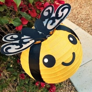 May include: A yellow and black bee-shaped paper lantern with a smiling face and black wings. The lantern is hanging from a branch of a bush with red flowers.