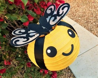 May include: A yellow and black bee-shaped paper lantern with a smiling face and black wings. The lantern is hanging from a branch of a bush with red flowers.