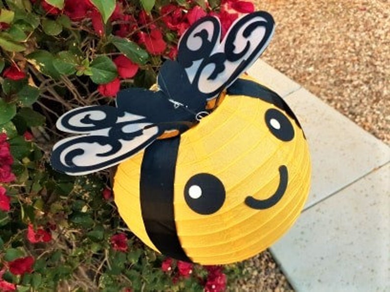 May include: A yellow and black bee-shaped paper lantern with a smiling face and black wings. The lantern is hanging from a branch of a bush with red flowers.
