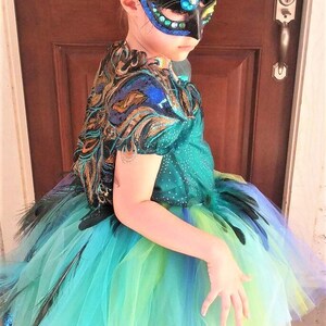 May include: A young person wearing a peacock-themed costume with a blue, green, and yellow tulle skirt, a black feather mask with rhinestones, and a green and gold sequined top.