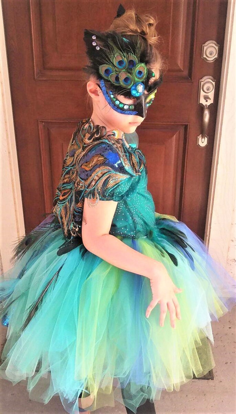 May include: A young person wearing a peacock-themed costume with a blue, green, and yellow tulle skirt, a black feather mask with rhinestones, and a green and gold sequined top.
