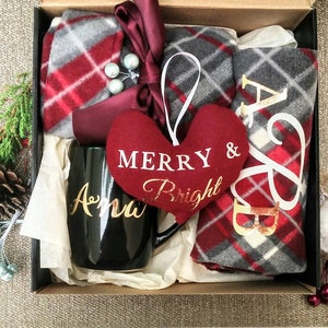 May include: A black gift box filled with a red and grey plaid blanket, a black mug with the name "Anna" in gold lettering, a red heart-shaped ornament with the text "Merry & Bright" in white, and a red and grey plaid blanket with the letters "A & B" in gold lettering.