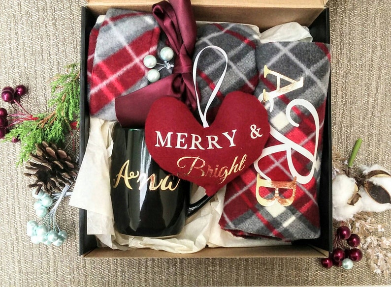 May include: A black gift box filled with a red and grey plaid blanket, a black mug with the name "Anna" in gold lettering, a red heart-shaped ornament with the text "Merry & Bright" in white, and a red and grey plaid blanket with the letters "A & B" in gold lettering.
