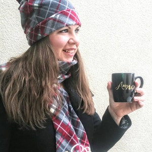 May include: A woman wearing a grey and red plaid hat and scarf with a matching pattern. She is holding a black mug with the word "Amo" written in gold lettering.