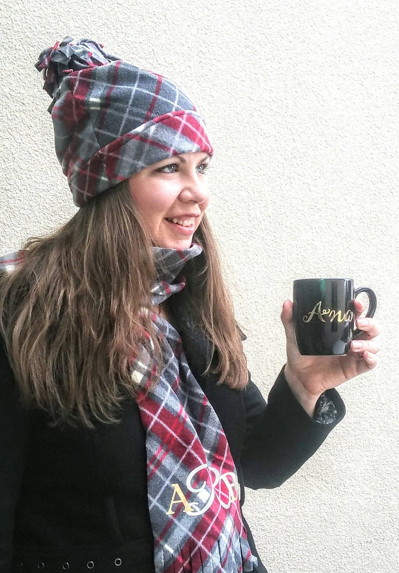 May include: A woman wearing a grey and red plaid hat and scarf with a matching pattern. She is holding a black mug with the word "Amo" written in gold lettering.