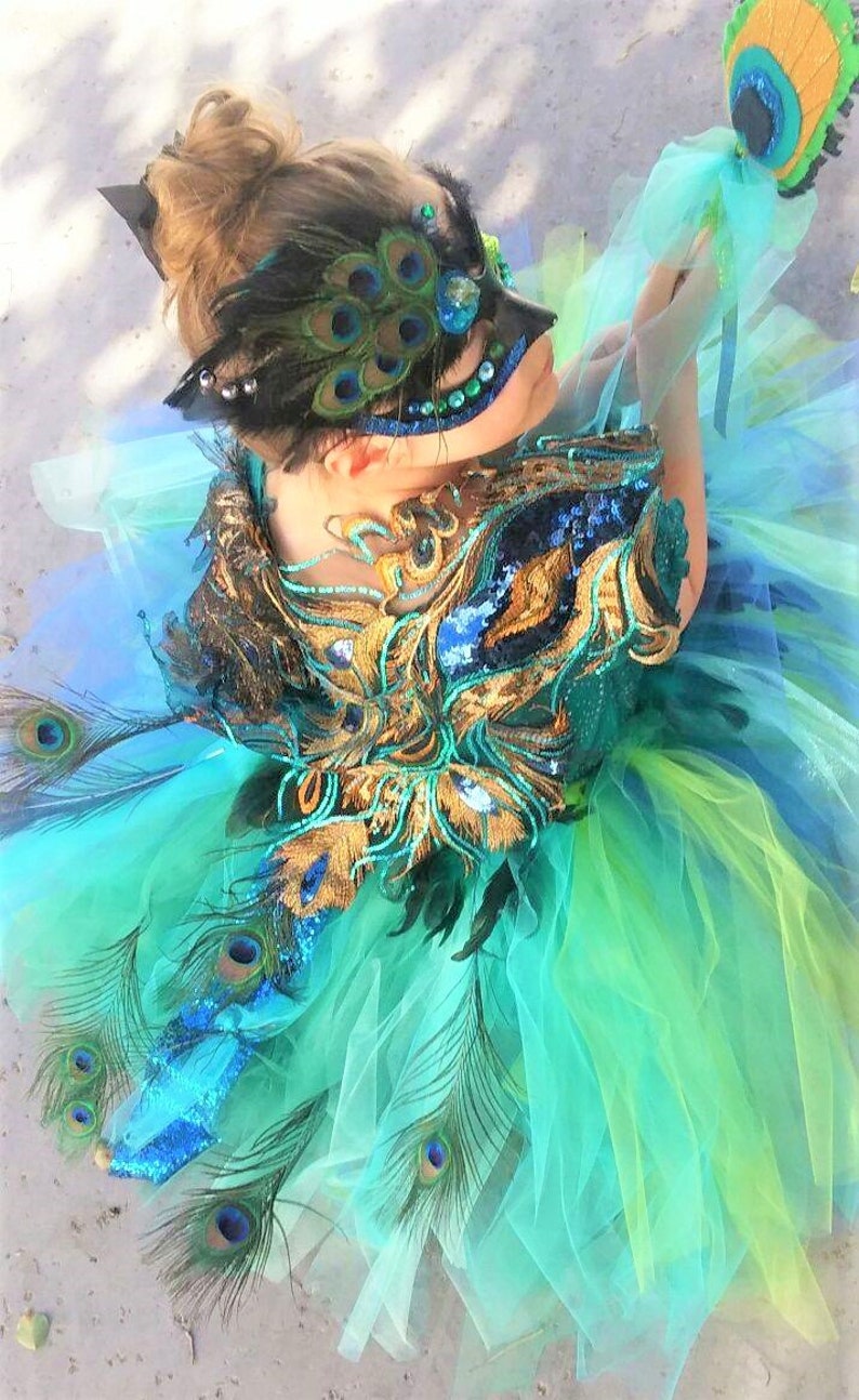May include: A girl wearing a peacock-themed costume with a blue and green tulle skirt, gold and blue sequined bodice, and a peacock feather mask.