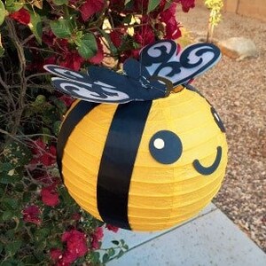 May include: A yellow and black paper lantern shaped like a bee with black wings and a smiling face.