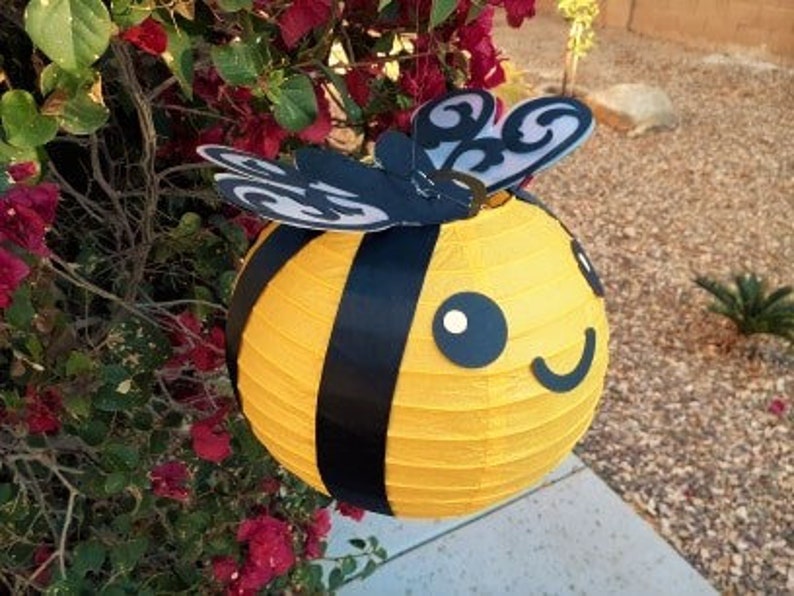 May include: A yellow and black paper lantern shaped like a bee with black wings and a smiling face.
