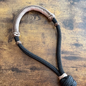 May include: A black and tan rope halter with a braided top section in shades of pink, tan, and brown. The halter features a black rope with a knotted end. The item is designed for equine use.
