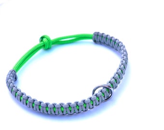 May include: A paracord bracelet or collar featuring a woven pattern of grey cord over a bright lime green core. The design includes a sliding knot closure for adjustment and a single circular metal ring integrated into the braided section.