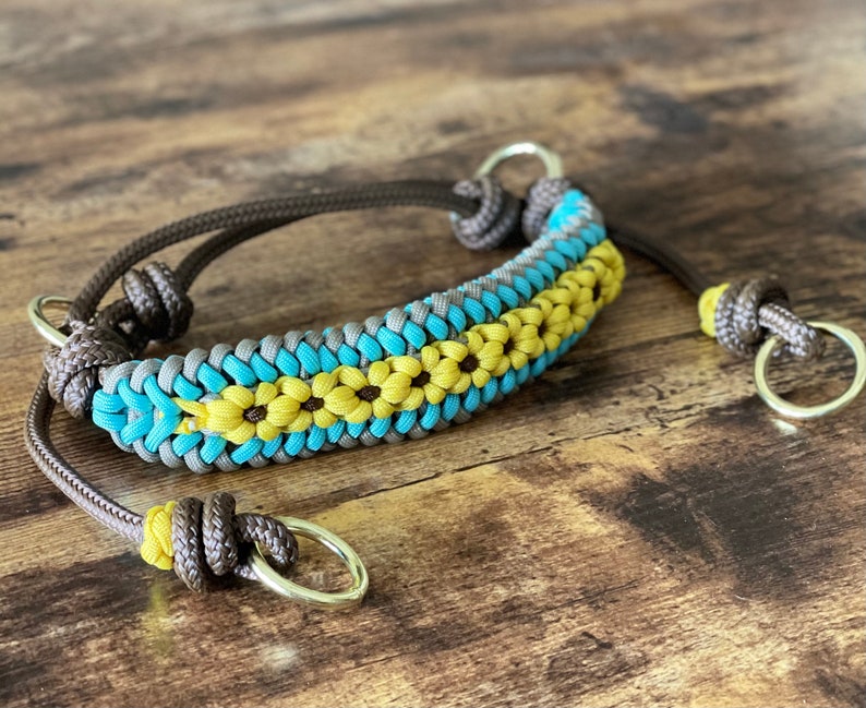 Pu&ograve; includere: Un collare per cani in paracord marrone con un design intrecciato blu e giallo con una fila di fiori gialli. Il collare ha due anelli metallici argentati.
