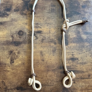 Puede incluir: Un cabestro de caballo de cuerda beige con detalles de cuero marrón. El cabestro tiene nudos y bucles para ajuste y fijación. La cuerda es de color beige claro y los detalles de cuero son de color marrón oscuro.