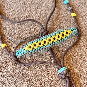Op de afbeelding: Een bruin touwhalster met een neusriem gevlochten in een zonnebloempatroon van geel, bruin en turquoise paracord. Het halster is voorzien van extra turquoise, grijze en gele knoopaccenten langs de zijriemen en een lus aan de onderkant.