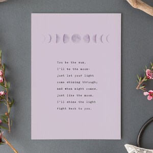 May include: A lavender print featuring a moon phase design at the top and a poem in a typewriter font. The poem reads, "You be the sun, I'll be the moon..." The print is on a gray background with floral accents.