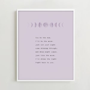 May include: A framed print featuring a light purple background with a series of moon phase illustrations. The print includes the text "You be the sun, I'll be the moon..." in a typewriter font, creating a celestial theme.