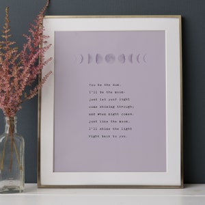 May include: Framed art print with a light purple background, featuring a moon phase graphic and a poem in black text. The frame is silver. A vase of pink flowers sits to the left.