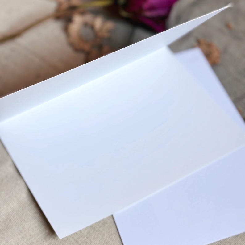 May include: A blank white greeting card, open to reveal the inside. The card is made of thick, bright white paper. The top flap is slightly raised, showing the interior. The card is on a neutral-toned surface.
