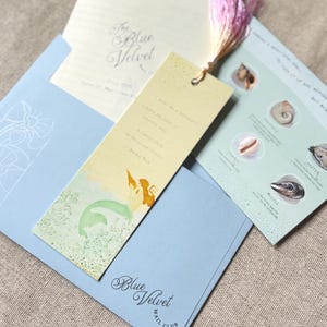 May include: A light blue envelope with the text Blue Velvet Mail Club, containing a yellow bookmark with a mermaid illustration and a quote by Anais Nin, a card with shell graphics, and a paper insert with a pink tassel attached to the top.