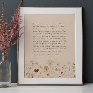 May include: A framed print featuring a quote from "The Curious Case of Benjamin Button." The text is on a beige background with floral illustrations. The frame is gold-coloured.