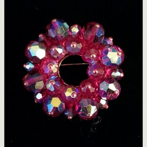 Op de afbeelding: Een roze en iriserende broche in de vorm van een cirkel. De broche is gemaakt van veel kleine, gefacetteerde kralen.