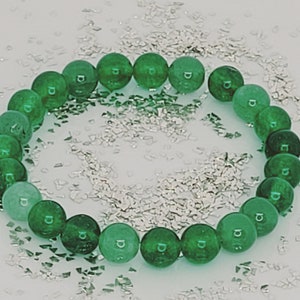 Peut inclure: Un bracelet en pierres précieuses vertes avec des perles rondes. Le bracelet est posé sur une surface blanche avec des paillettes argentées.