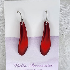 May include: A pair of red dangle earrings with silver hooks. The earrings are shaped like a teardrop and have a shiny, metallic finish. The earrings are displayed on a white card with the text "Bella Accessories By Julie Misasi Made in Canada".