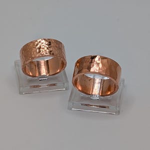 May include: Two wide copper rings featuring a hammered, textured surface. Each ring has a polished metallic finish and a consistent band width, displayed upright on individual clear square stands to showcase the circular shape and reflective metal.