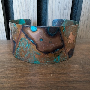 May include: A wide copper cuff bracelet with a distinctive patina. The bracelet displays shades of brown, teal, and blue, creating a textured, artistic look. The open design allows for easy wear. A statement piece of jewellery.
