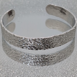 May include: A silver-toned metal cuff bracelet featuring a textured, hammered surface with an uneven, organic pattern. The bracelet is open-ended, forming a C-shape, and has a reflective, metallic finish that catches light across its entire exterior.