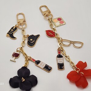 May include: Two gold keychains with various charms. One keychain has a black cowboy hat, a black boot, a wine glass, a bottle of wine, and a black flower. The other keychain has a red lipstick, a pair of glasses, a bottle of wine, and a red flower.