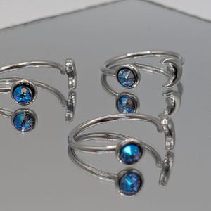 Peut inclure: Trois bagues argentées avec un motif de croissant de lune et une pierre précieuse bleue. Les bagues sont présentées sur une surface réfléchissante. Les pierres précieuses ont un aspect facetté.