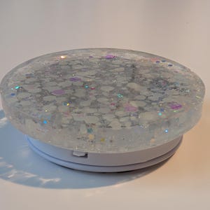 May include: A circular phone grip featuring a clear resin top embedded with white, iridescent, and holographic glitter flakes. The base is a solid white plastic collapsible mechanism that allows for expansion and retraction when attached to a device.