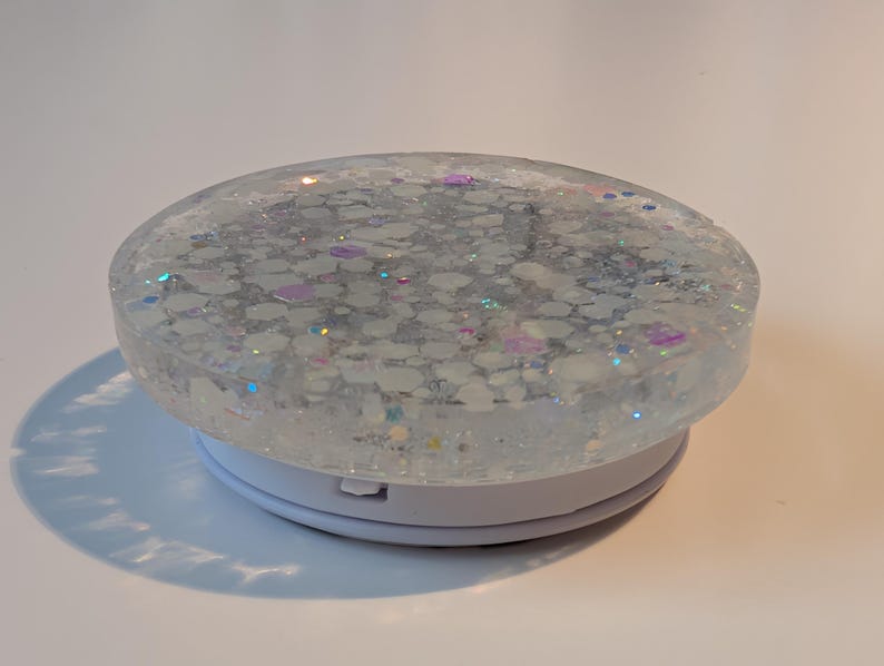 May include: A circular phone grip featuring a clear resin top embedded with white, iridescent, and holographic glitter flakes. The base is a solid white plastic collapsible mechanism that allows for expansion and retraction when attached to a device.