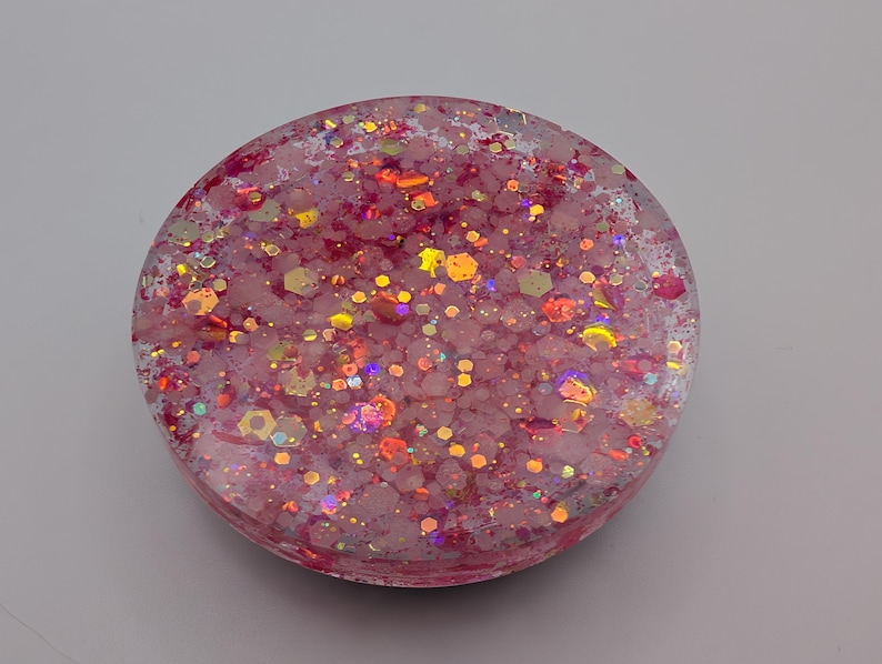May include: A circular resin coaster featuring a translucent pink base embedded with a dense mixture of multi-sized hexagonal and fine glitter particles in shades of pink, gold, and iridescent orange that reflect light across the surface.