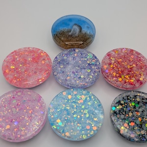 Könnte beinhalten: Sieben kreisförmige Handygriffe aus Harz, die in einem Dreieck angeordnet sind. Einer zeigt ein blau-braunes Design, das an einen Strand erinnert, während die anderen sechs mit verschiedenen bunten, reflektierenden und holografischen Glitzerpartikeln in Rosa, Lila, Blau und Schwarz gefüllt sind.