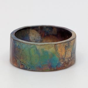 May include: A wide band ring featuring a multi-coloured, iridescent finish with mottled patches of blue, green, bronze, and dark brown. The surface has a textured, hammered appearance with a slightly uneven edge, creating a distinct metallic look.